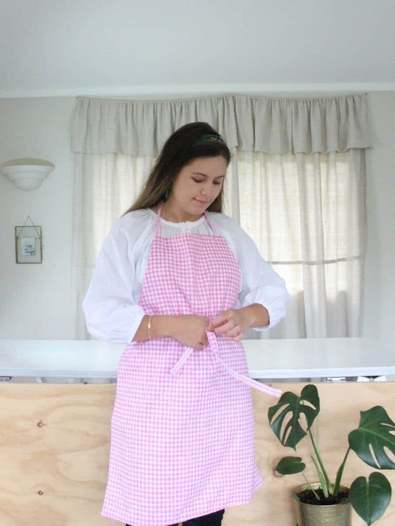 How To Make A Apron: Step By Step makyla is wearing a pink gingham apron while tying her waist tie into a bow at the front