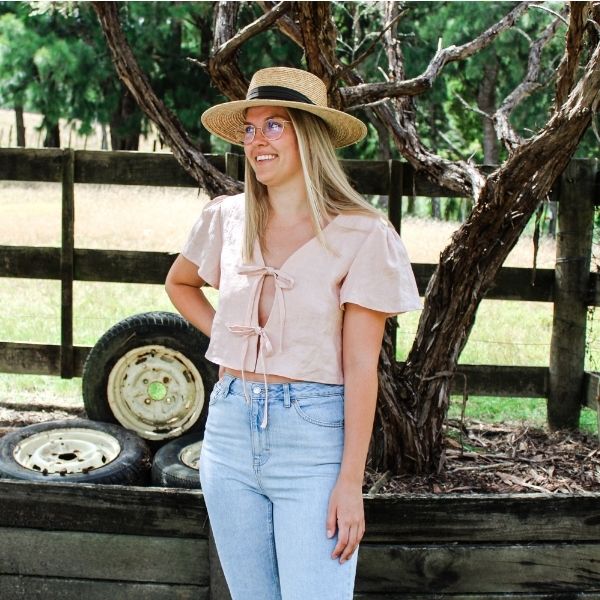 Shania wearing pink linen camille top with front ties and flute sleeves. She is smiling and wearing a straw hat. There are paddocks and fences behind her.