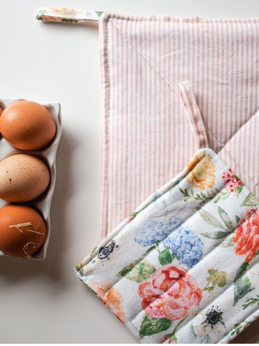 White and floral pot holder with stripe tab laying on white bench top. There is a white/pink striped potholder behind it on a white bench. There are eggs in the corner of the shot that are sitting in a ceramic egg tray.