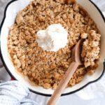 Apple crumble cooked in a red cast iron pan. A scoop of vanilla ice cream is on top with a wooden spoon to serve