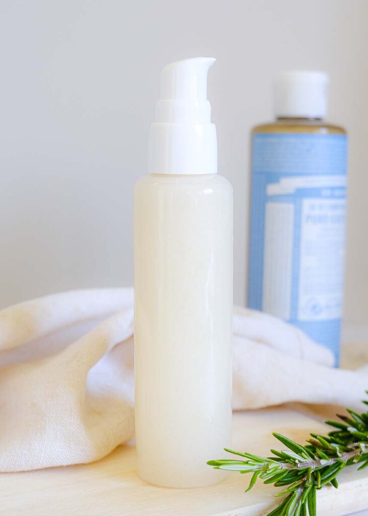 Homemade shampoo poured into a small clear bottle with spuirt nozzle. There is fresh rosemary sprigs and a bottle of unscented castile soap in the background.