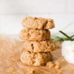 Four anzac biscuits stacked on top of each other. They are sitting on a wooden chopping board with brown parchment paper.