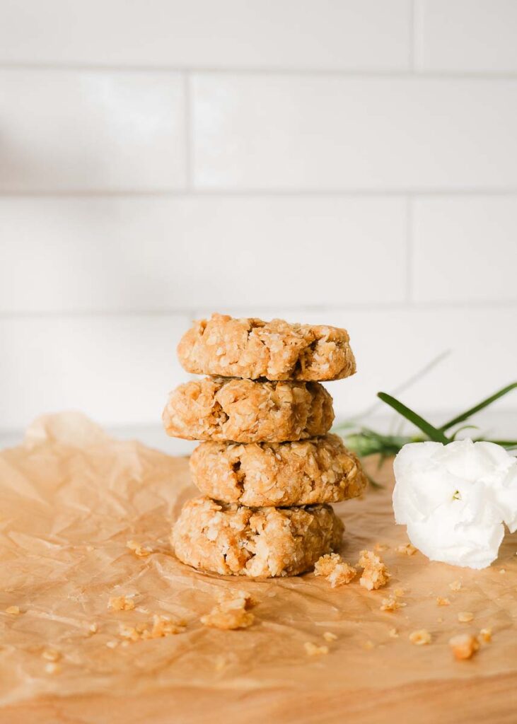 Four freshly baked anzac biscuits stacked in a pile on a wooden chopping board next to fresh white flowers. There are cookie crumbs all over the board