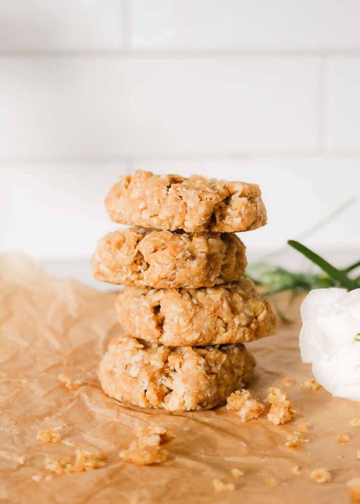 Four freshly baked anzac biscuits stacked in a pile on a wooden chopping board next to fresh white flowers.