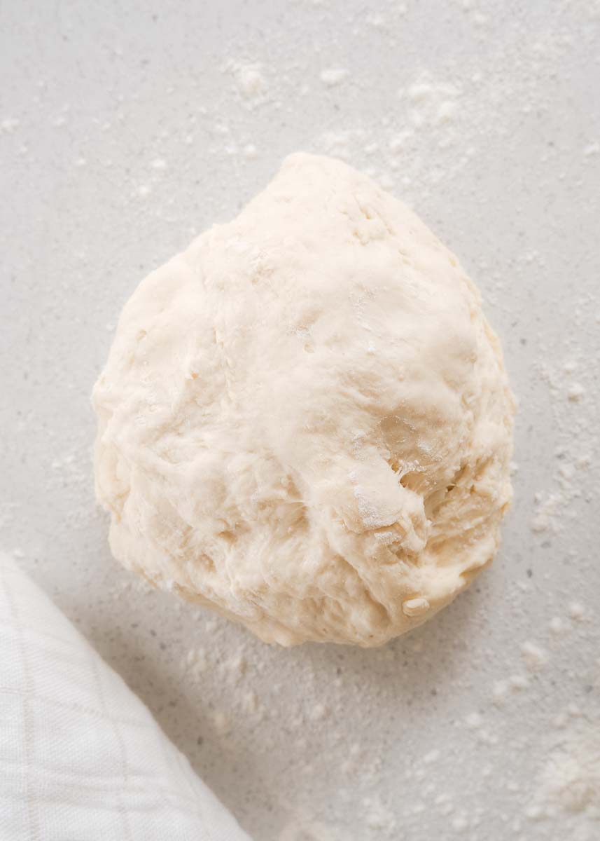 The ball of dough on the kitchen bench which is covered in flour