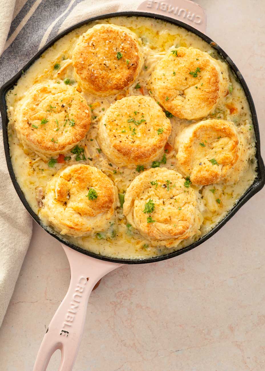 Golden baked biscuits on top of the creamy chicken pot pie filling in a pink-handled skillet, garnished with herbs.