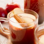 A close-up of a mug of apple cider with floating apple slices and a cinnamon stick, with a jar of cider and red apples in the background.