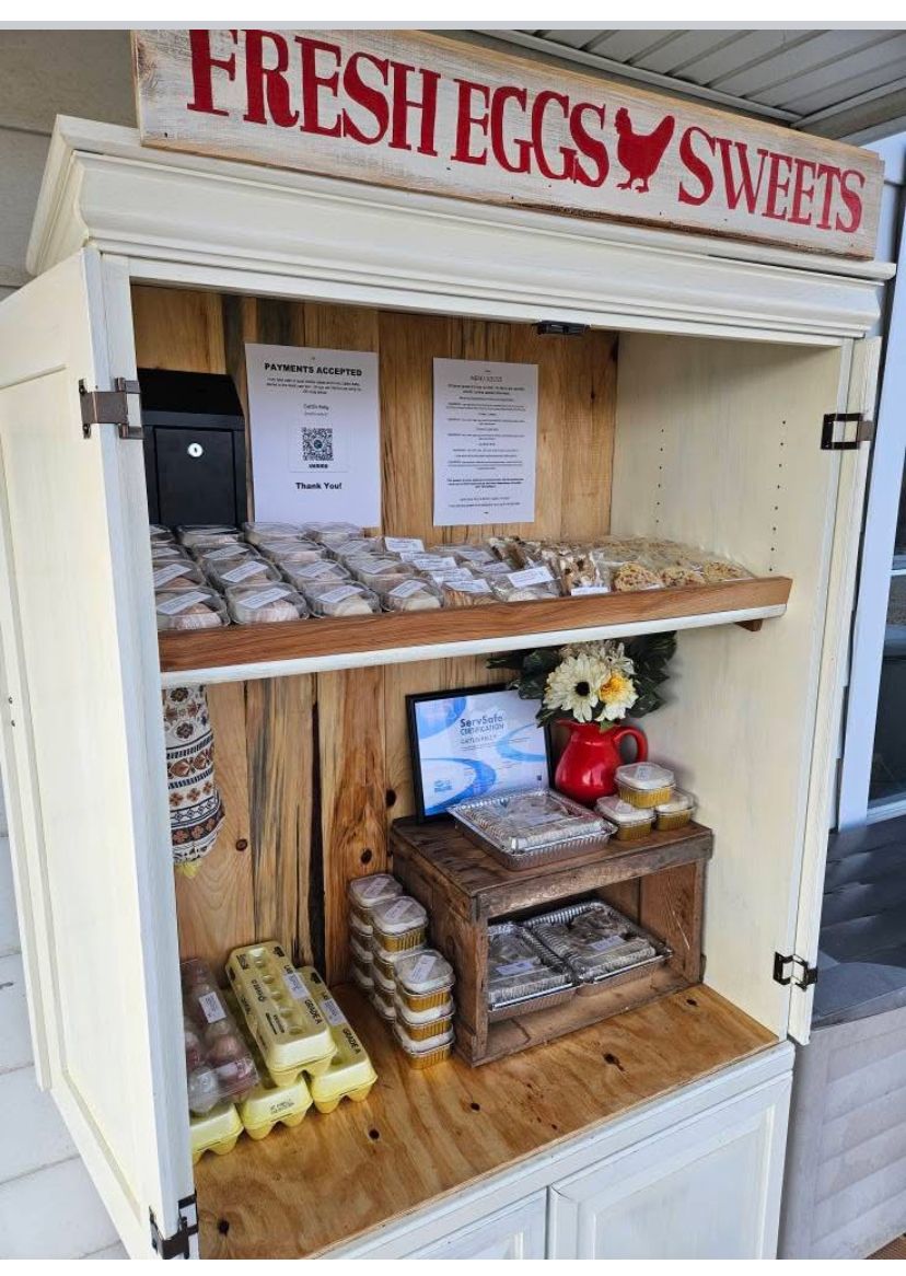 Cabinet-style farmstand with &ldquo;Fresh Eggs & Sweets&rdquo; sign, eggs, baked treats, and flowers.