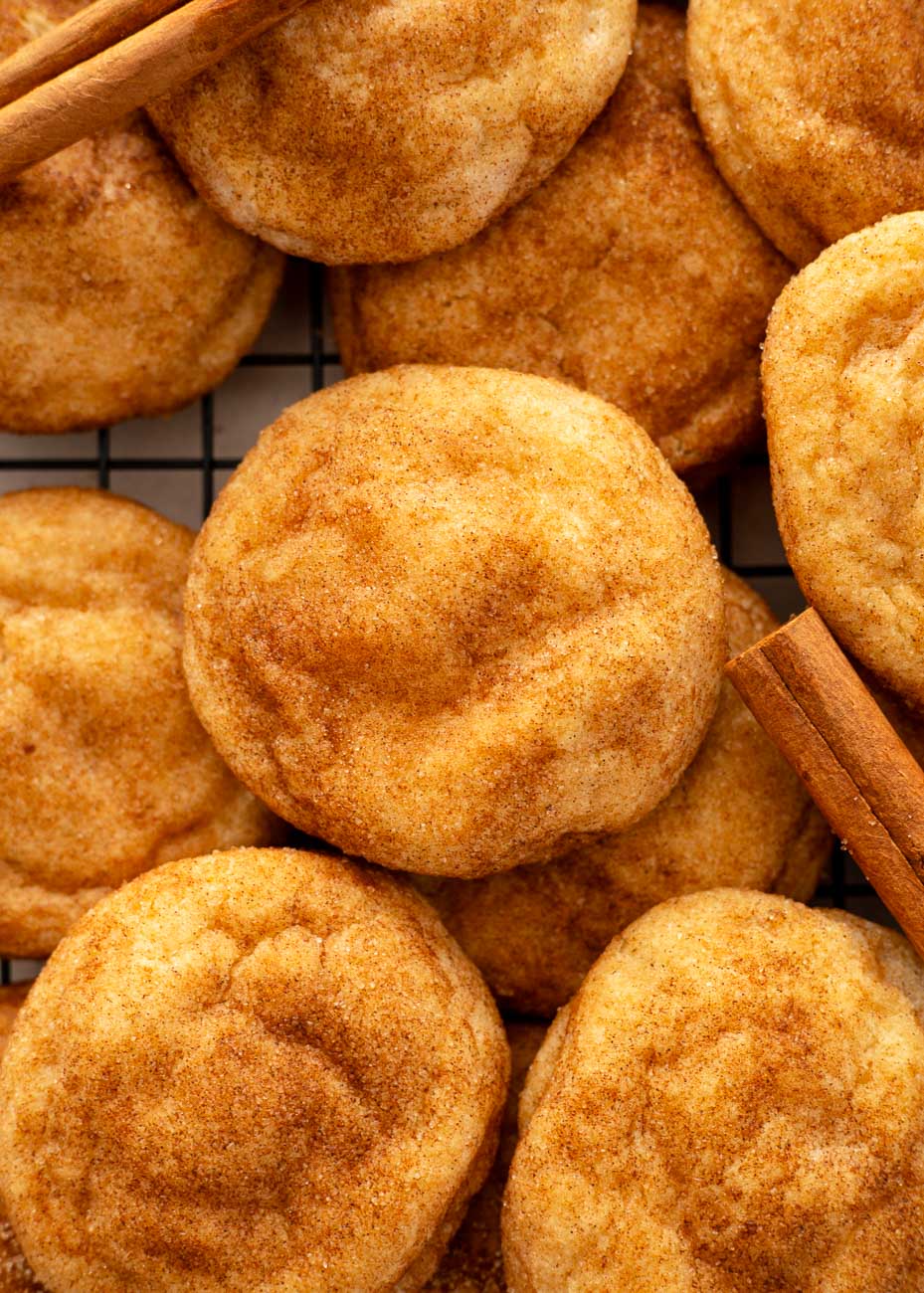 A close-up shot of a pile of golden-brown snickerdoodle cookies on a wire rack, sprinkled with cinnamon sugar.