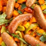 Tight close-up of a cooked, glazed sausage on a tray bake of roasted orange pumpkin and sweet potato, green beans, and bell peppers, garnished with fresh thyme.