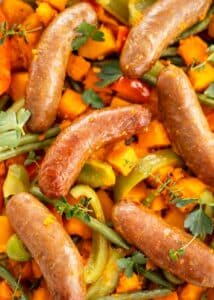 Tight close-up of a cooked, glazed sausage on a tray bake of roasted orange pumpkin and sweet potato, green beans, and bell peppers, garnished with fresh thyme.
