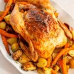The whole roast chicken served on a white platter, resting on a bed of golden roasted potatoes and carrots, garnished with fresh herbs.