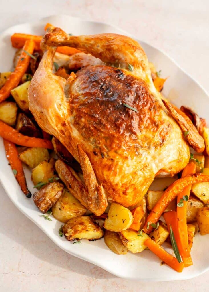 The whole roast chicken served on a white platter, resting on a bed of golden roasted potatoes and carrots, garnished with fresh herbs.