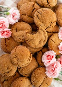 Close-up of a pile of freshly baked, sugar-coated molasses cookies with crackled tops, surrounded by pink carnations. One cookie has a bite taken out of it.