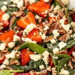 A close-up, high-angle shot of the assembled strawberry spinach salad, showing the detail of the sliced strawberries sprinkled with poppyseeds, crumbled feta, chopped pecans, and spinach leaves.