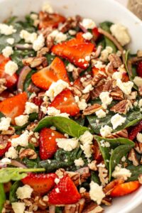 A close-up, high-angle shot of the assembled strawberry spinach salad, showing the detail of the sliced strawberries sprinkled with poppyseeds, crumbled feta, chopped pecans, and spinach leaves.