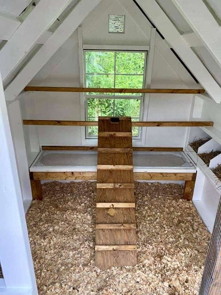 Bright white-painted A-frame chicken coop interior with staggered horizontal timber roosting bars spanning the full width, large picture window overlooking green garden, central wooden ladder-style ramp, and wood chip covered floor. Nesting area visible on the right.