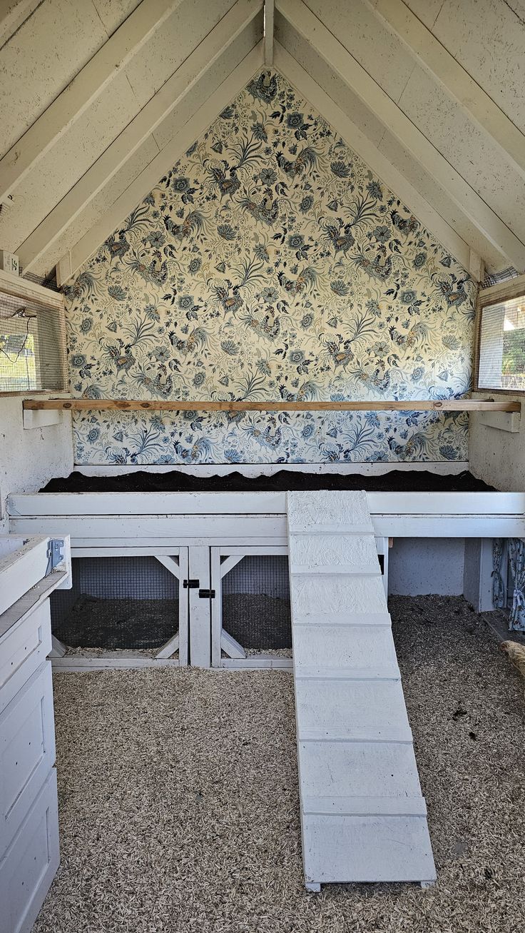 Charming A-frame chicken coop interior with white-painted timber throughout, blue and cream toile de Jouy rooster and floral pattern wallpaper covering the full gable end wall, wide raised droppings board, cabinet-style nesting boxes with wire mesh doors underneath, and white painted ramp with grip strips leading up to the roosting level.