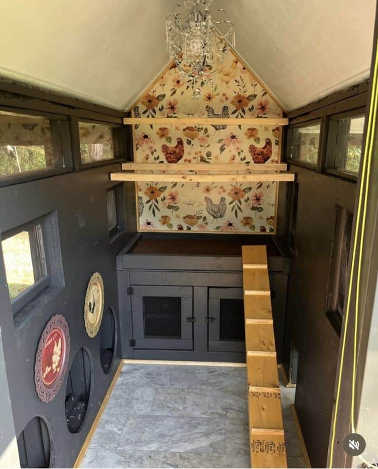 Dramatic dark charcoal-painted A-frame chicken coop interior with crystal chandelier hanging from the vaulted ceiling, warm floral wallpaper with chicken motifs covering the gable feature wall, staggered timber roosting bars, dark painted nesting cabinet with panelled doors, slate tile flooring, decorative rooster plate on the side wall, and timber ramp leading up to the roost.