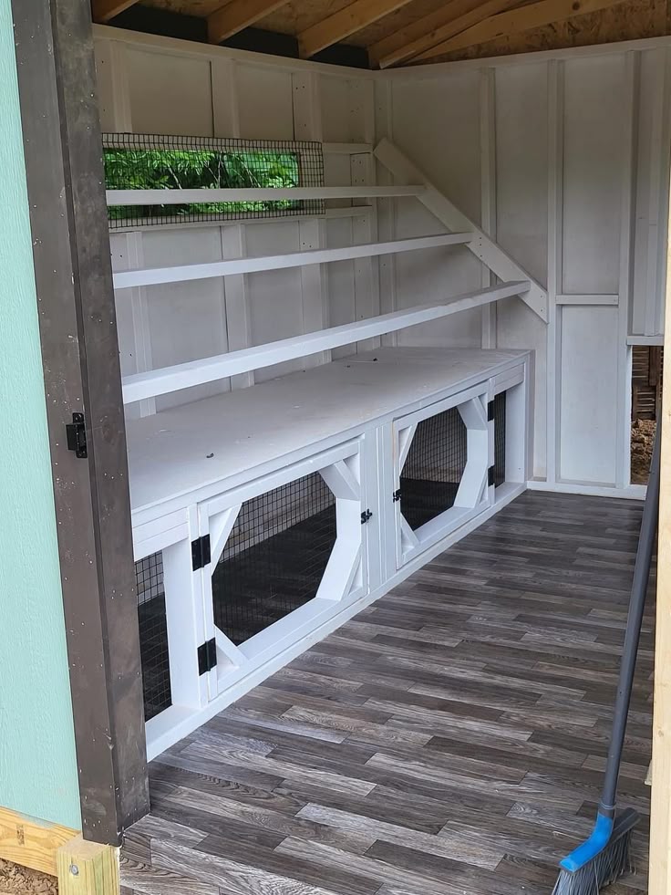 Clean, elegant chicken coop interior with white-painted walls, custom built-in nesting cabinet with arched wire mesh doors, staggered white roosting bars above, and grey vinyl plank flooring