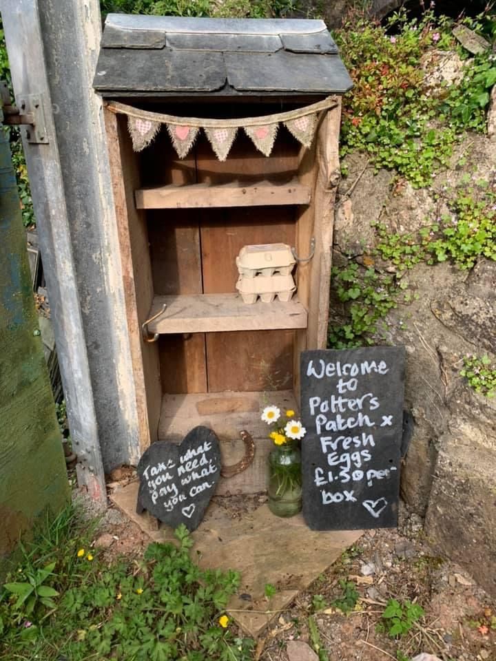 A small rustic open wooden cabinet egg stand made from aged and weathered timber, decorated with hessian bunting trimmed with pink heart patches, a single egg carton on the middle shelf, and two handwritten chalkboard signs leaning beside it, one reading Welcome to Potters Patch Fresh Eggs and one reading Take What You Need Pay What You Can