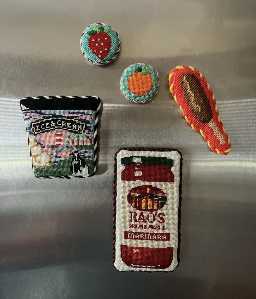 Five stuffed needlepoint fridge magnets on a stainless steel fridge including a Rao&rsquo;s marinara jar, an ice cream pint, a hot dog, a strawberry, and an orange