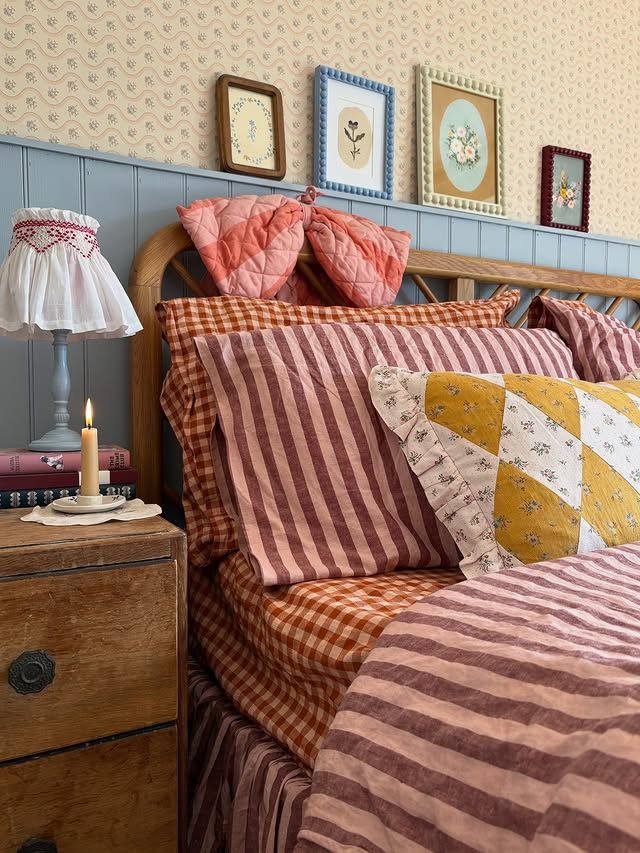 Cottagecore bedroom with gingham and striped bedding in burgundy and terracotta, smocked lamp, and framed botanical wall art above a wooden headboard