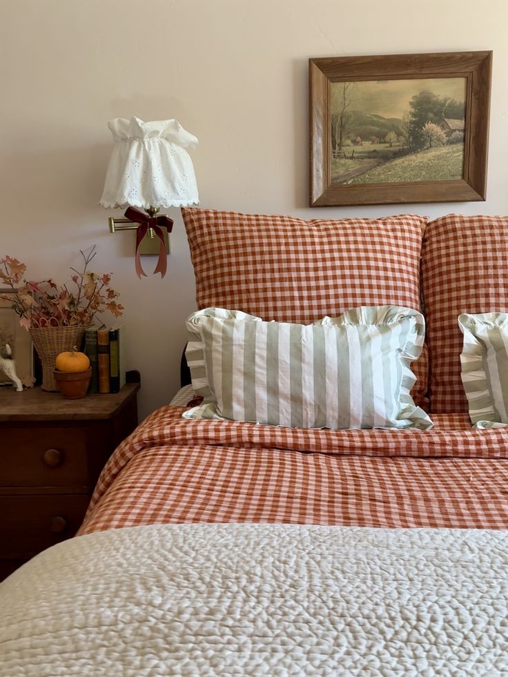 Cottagecore bedroom with terracotta gingham duvet and pillow shams, sage green ticking stripe lumbar pillow, white eyelet wall sconce, framed landscape art, and an autumn-styled wooden nightstand