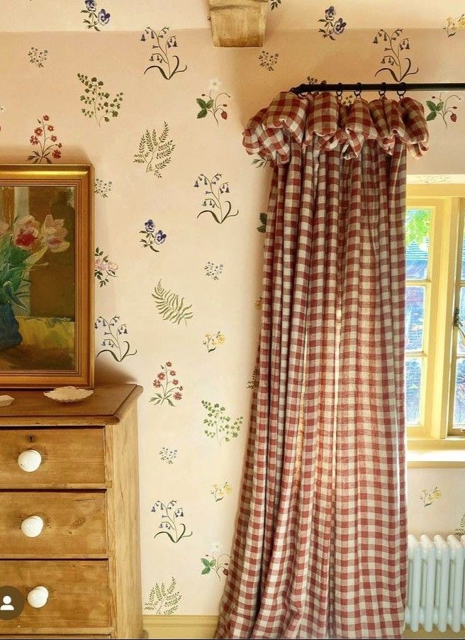 Cottagecore bedroom with hand-painted wildflower and botanical motifs on white walls, deep red gingham curtains with ruffled tab tops, a yellow window frame, and a pine chest of drawers with ceramic knobs