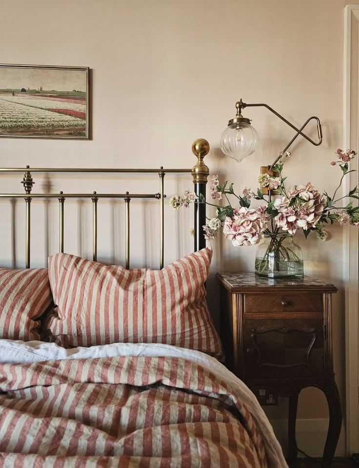 Romantic cottagecore bedroom with a brass antique-style metal bed frame, wide stripe linen bedding in dusty pink and white, a carved wooden nightstand with pink flowers in a glass vase, and a brass swing arm wall sconce