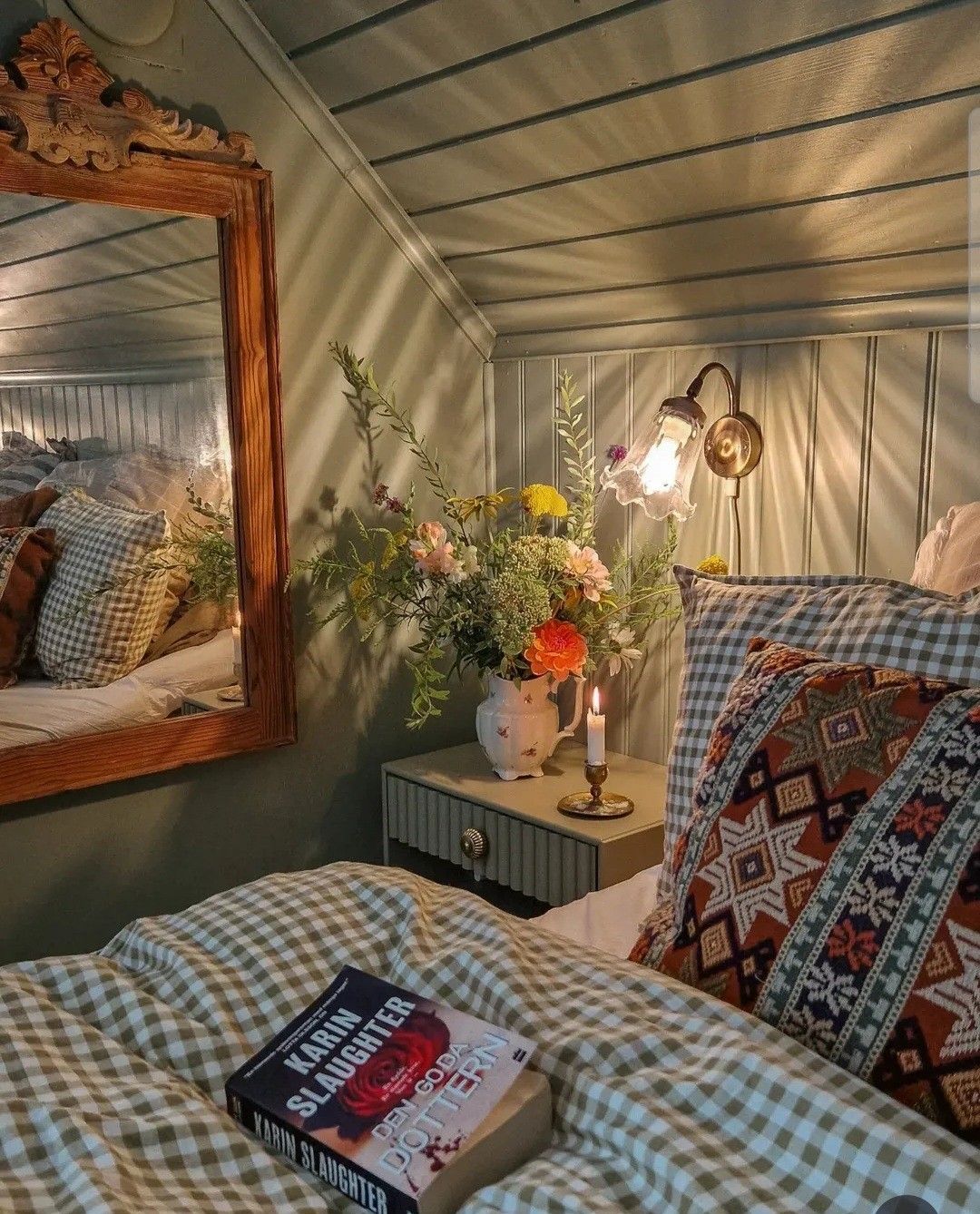 Cosy cottagecore bedroom with sage green tongue and groove panelled walls, gingham bedding, a wildflower arrangement in a china pitcher on the nightstand, a brass candle holder, and a carved wood-framed mirror