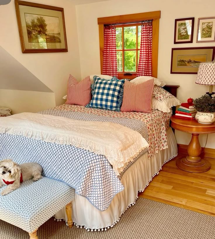 Cottagecore country bedroom with red gingham window curtains, ticking stripe and navy gingham pillows, floral quilt, ruffled bed skirt, a pom pom trim bench at the foot of the bed, and framed landscape prints