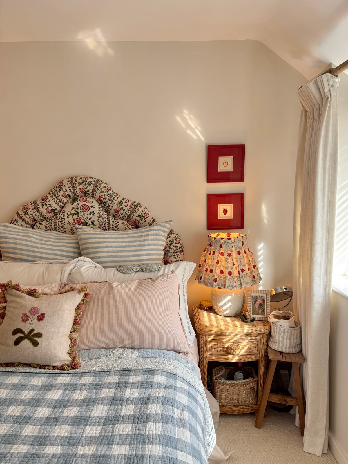 Cottagecore bedroom with a cream floral scalloped upholstered headboard, blue gingham quilted bedding, an embroidered tassel cushion, a rattan nightstand with a block print lamp shade, and red-framed strawberry botanical art