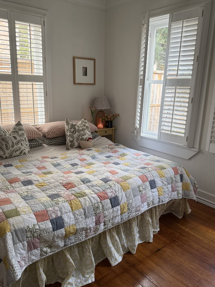 Cottagecore bedroom with a multicolour patchwork quilt in pastel prints, a ruffled floral bed skirt, plantation shutters on double windows, a small nightstand with fresh flowers, and paisley and gingham accent pillows