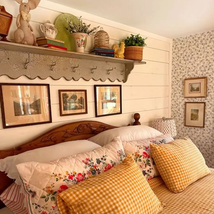 Cottagecore bedroom with a white shiplap feature wall, a vintage scallop-edged coat rack shelf styled with ceramics and a wicker basket, three framed landscape prints above a carved wooden headboard, and yellow gingham bedding