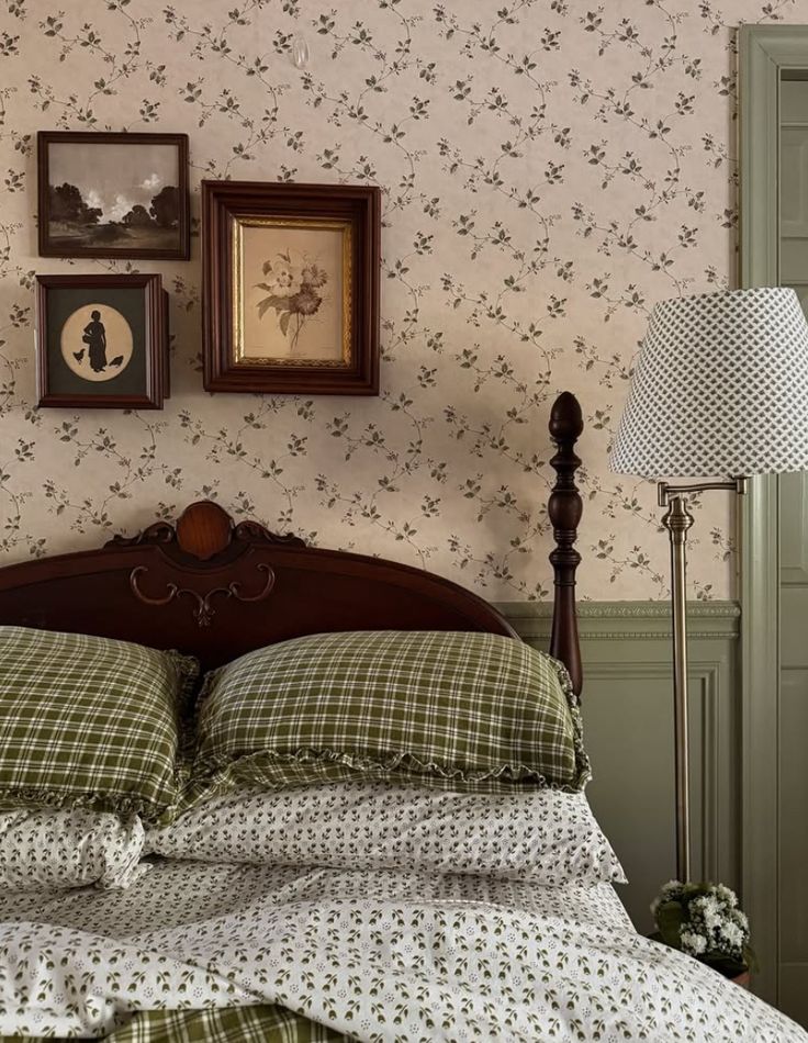 Romantic cottagecore bedroom with trailing green floral wallpaper, dark wood antique bed frame, green tartan ruffled bedding, and a gallery wall of framed artwork