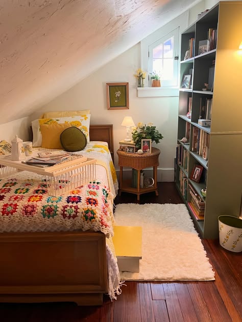 Cottagecore bedroom with a multicolour granny square crochet throw over white bedding, yellow floral pillow shams, a round wicker nightstand, a green bookcase filled with books and plants, and a breakfast tray on the bed