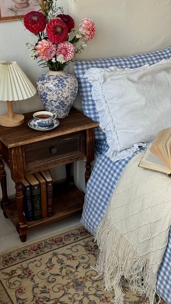Cottagecore bedroom nightstand with a blue and white chinoiserie vase filled with pink and red dahlias, a pleated lamp, teacup, and a stack of books, beside blue gingham bedding