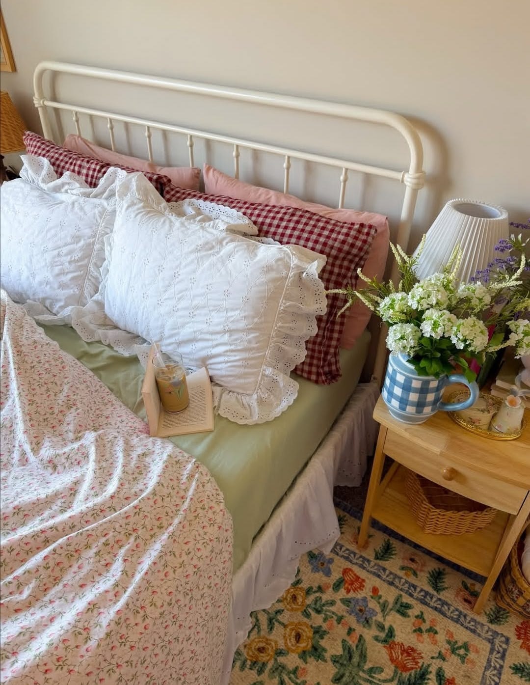 Cottagecore bedroom with a cream iron bed frame, white eyelet pillow shams, red gingham and pink pillows, a floral quilt, and a blue gingham mug with wildflowers on the nightstand