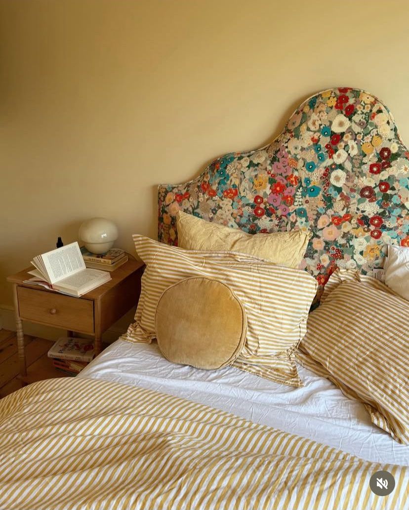 Cottagecore bedroom with a bold colourful floral fabric upholstered headboard, golden yellow stripe duvet and pillow shams, a round velvet cushion, and warm ochre walls