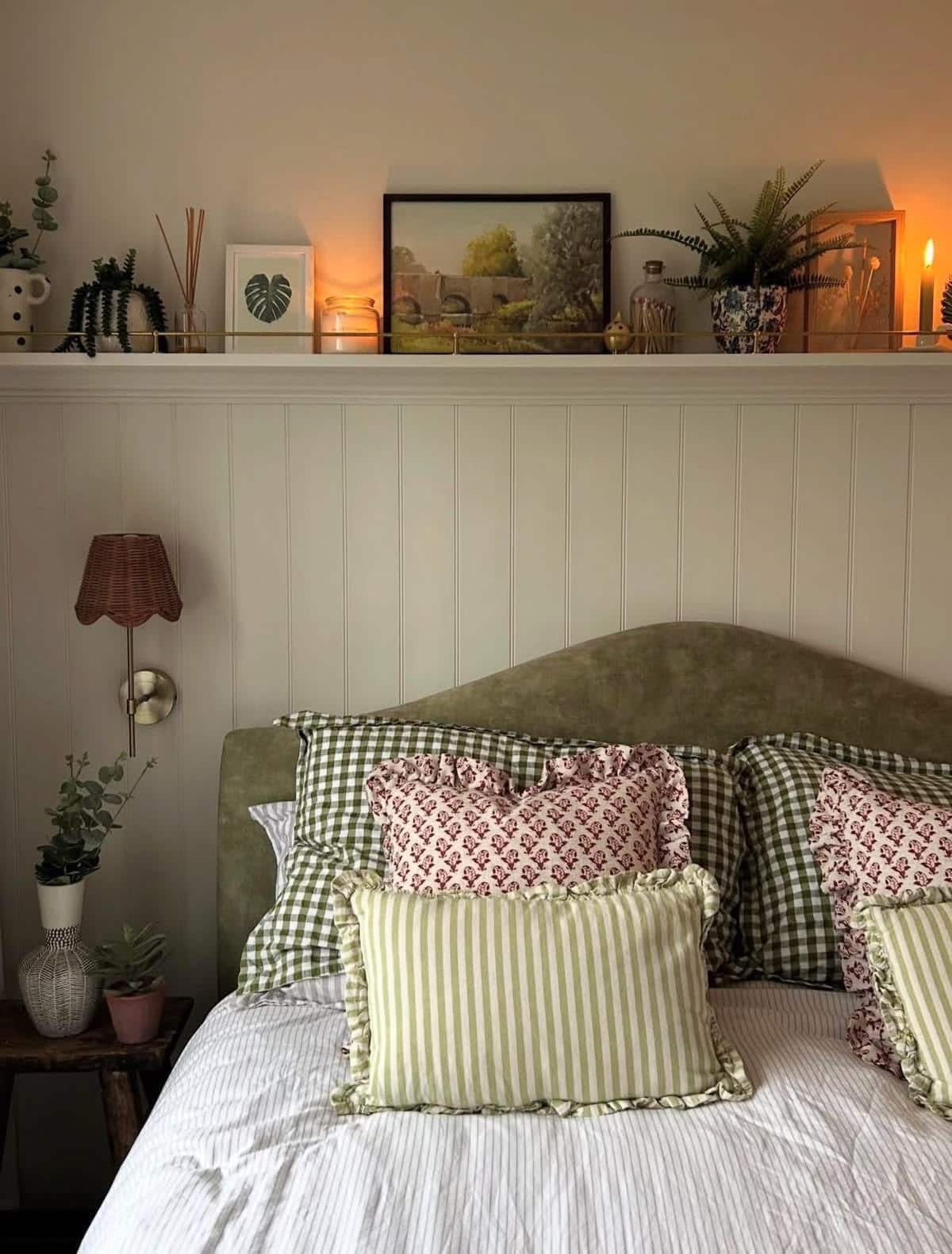 Cottagecore bedroom with white shiplap wall, a display shelf above the bed styled with candles and plants, an olive velvet headboard, green gingham bedding, and a rattan wall sconce