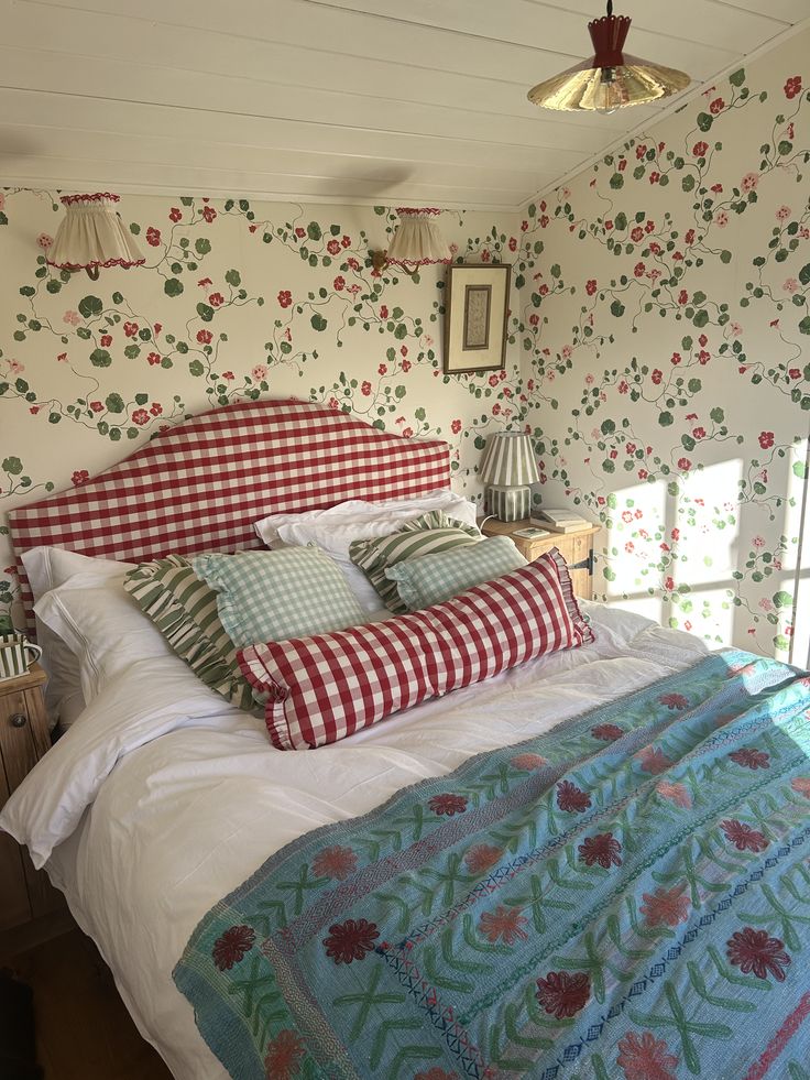 Bold cottagecore bedroom with red and green floral nasturtium wallpaper, red gingham upholstered headboard, white bedding, gingham and ticking stripe accent pillows, and an embroidered teal quilt