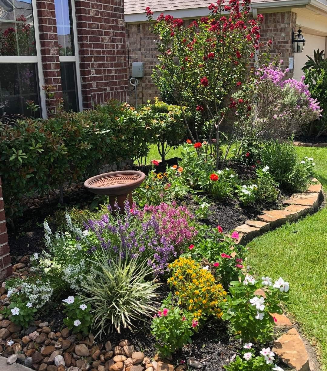 A lush layered front yard garden bed with a crape myrtle tree, bird bath, purple salvia, impatiens, and rudbeckia flowers against a brick home exterior with stone edging.