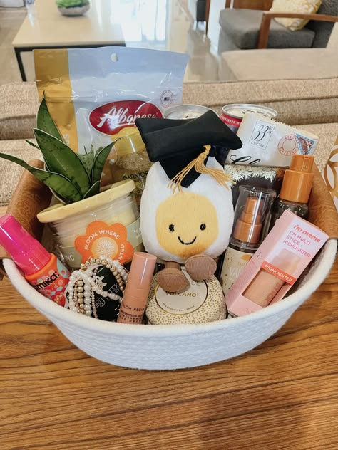 A white rope bowl filled with a small succulent plant, a Bloom Where You Are Planted candle, a happy egg plushie in a graduation cap, Pearl hair clips, a Volcano candle, I'm Meme highlighter, and golden body mist.
