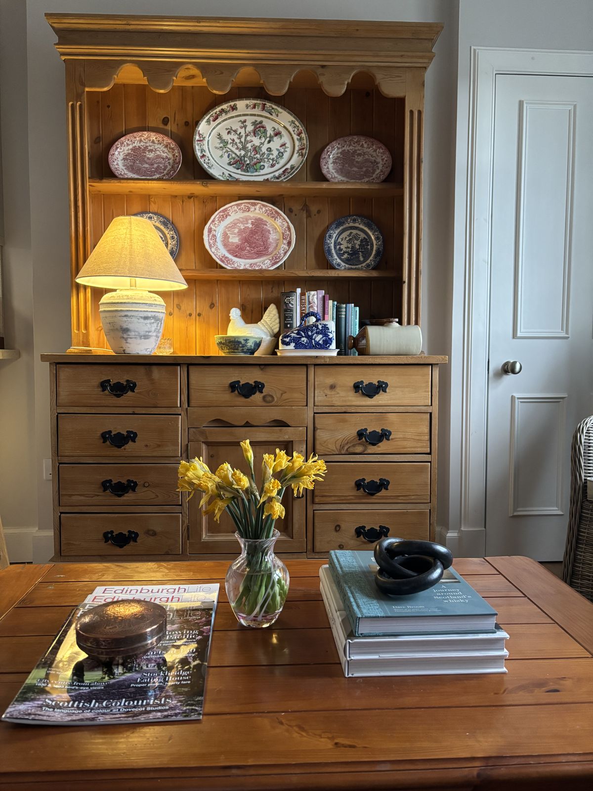 Pine hutch with red and blue transferware plates displayed on upper shelves a ceramic lamp and yellow daffodils in a glass vase on the surface below