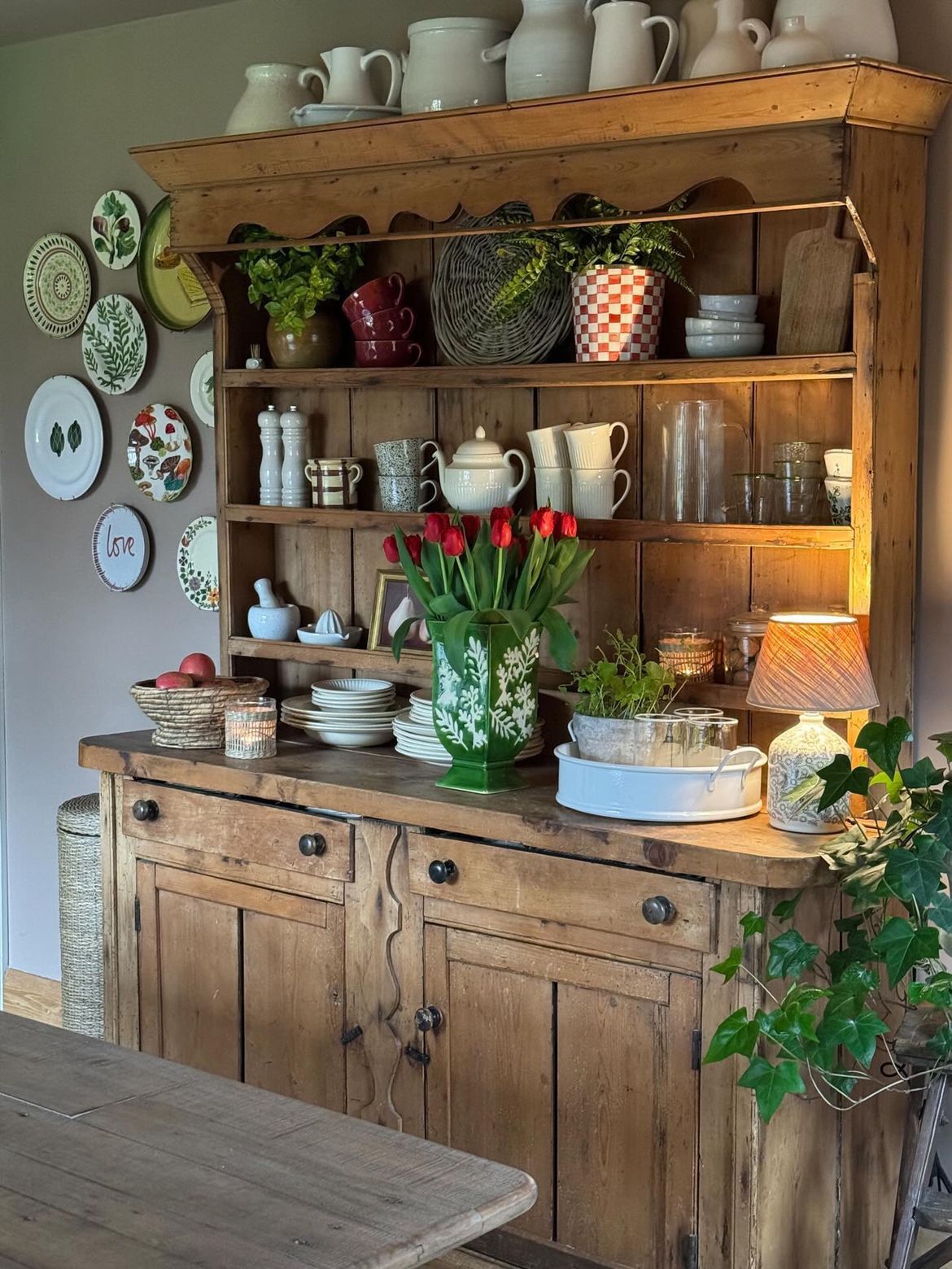 Large pine hutch with red tulips in a green painted vase mixed white ceramics a warm table lamp and a plate gallery wall beside it