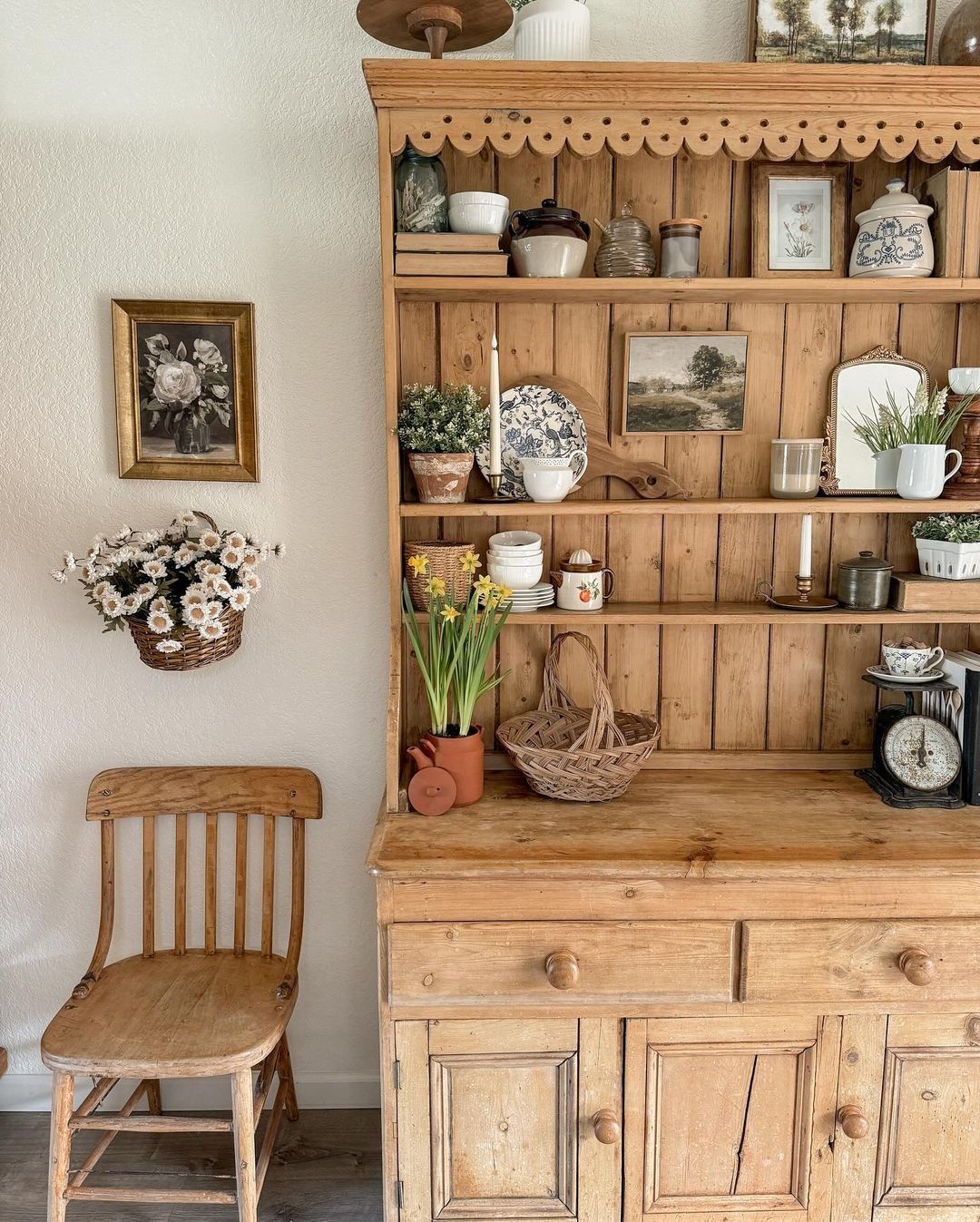 Pine hutch with a scalloped cornice styled with framed botanical prints candlesticks daffodil bulbs in a terracotta pot wicker baskets and vintage kitchen scales