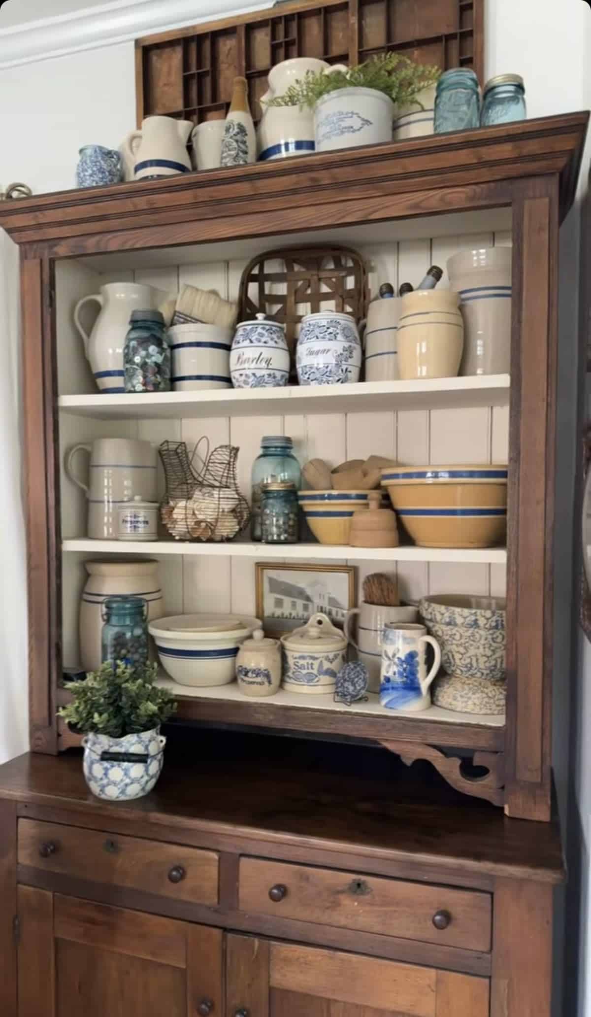 Dark pine hutch with white-painted shelves filled with antique stoneware crocks salt-glazed pottery yellow mixing bowls mason jars and a small framed print