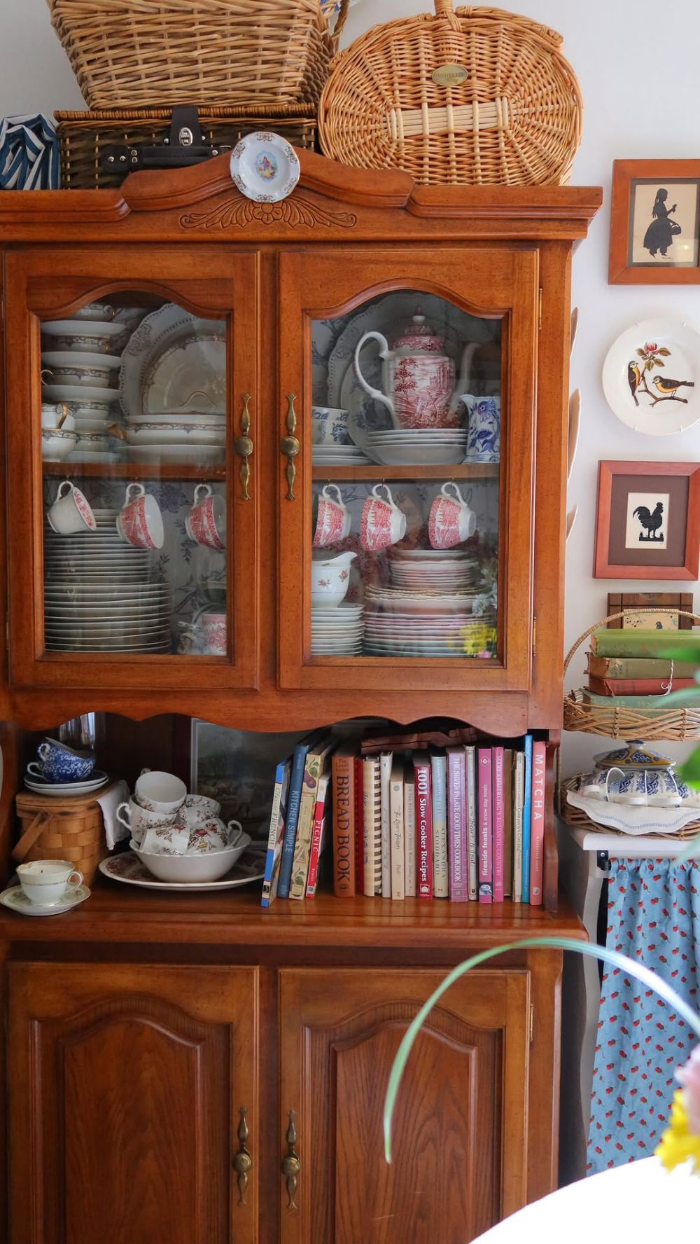 Glass-front timber hutch with red and white transferware tea sets plates and cups inside the glass doors and cookbooks and china on the open lower shelf