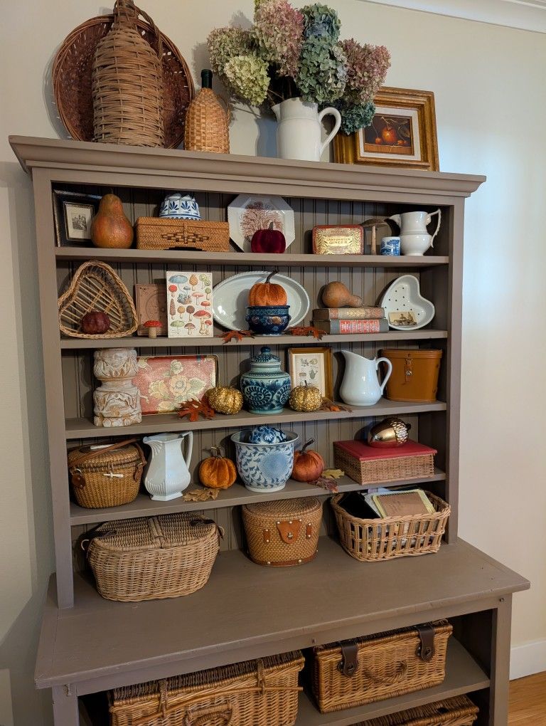 Greige painted hutch styled for autumn with velvet pumpkins dried hydrangeas blue and white ginger jars white pitchers wicker baskets and autumn leaf details